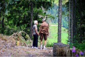 An older couple strolls hand in hand through a serene forest, surrounded by lush greenery and dappled sunlight.