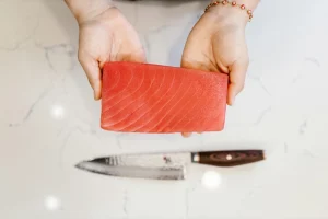 Both hands holding a slice of red fish meat and a knife is shown on the table below the hands.