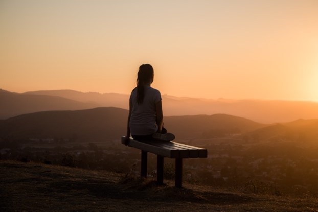 A girl sitting on a bench on top of a hill viewing the scenery up there.